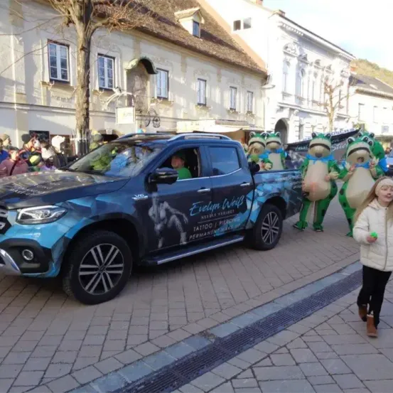 Ein blauer Pickup-Truck mit einer Person im Inneren fährt durch eine Parade, gefolgt von Menschen in Froschkostümen. Ein Kind geht auf dem Bürgersteig vor dem Truck.