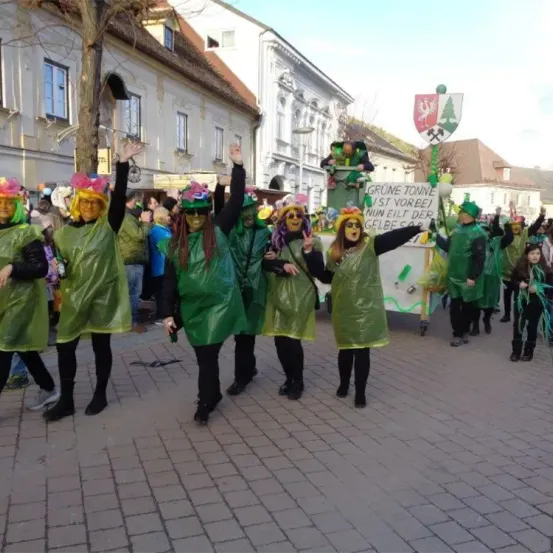 Eine Gruppe von Menschen in grünen Kostümen und Masken zieht durch einen Stadtplatz, hebt die Hände und lächelt, mit Gebäuden und einem geschmückten Wagen im Hintergrund.