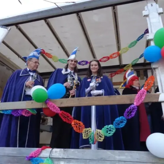 Drei Personen in blauen Kostümen und Hüten stehen auf einer Bühne mit Luftballons. Sie scheinen sich auf einer Feier mit mehrfarbigen Dekorationen zu befinden.