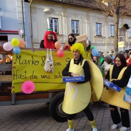 Drei Personen in gelben Kostümen, eine davon mit einer Box, stehen auf einem dekorierten Anhänger bei einer Parade. Dahinter steht eine Person in einem roten Kostüm auf dem Anhänger.