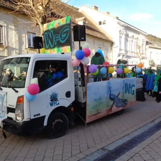 Ein weißer Lastwagen, dekoriert mit bunten Luftballons und einem Schild mit der Aufschrift W-G, steht auf der Straße. Eine Pfauenmalerei befindet sich an der Seite des Lastwagens.