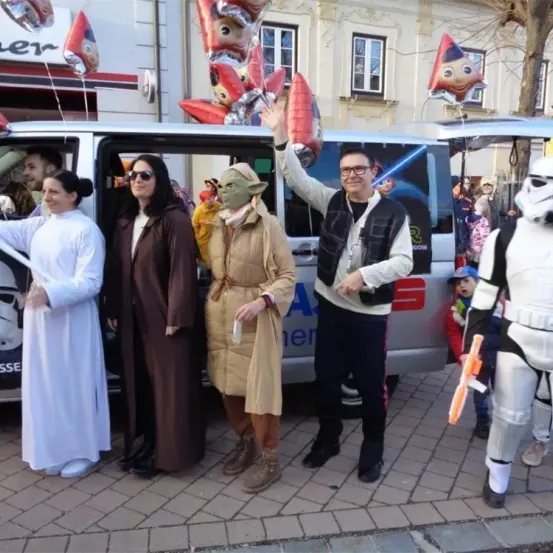 Fünf Menschen in Kostümen stehen neben einem Van, einer von ihnen winkt. Der Van hat Star Wars-Werbung an seinen Seiten. Hinter ihnen stehen weitere Menschen, einige in Sturmtruppler-Kostümen. Im Hintergrund sind Ballons und ein Baum zu sehen.