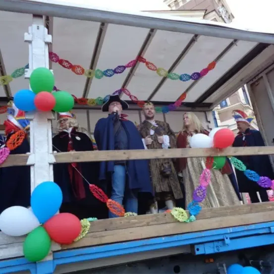 Eine Gruppe von Menschen in Kostümen steht auf einer dekorierten Plattform mit Luftballons, möglicherweise für eine Parade. Eine Person hält ein Mikrofon und scheint zu sprechen.