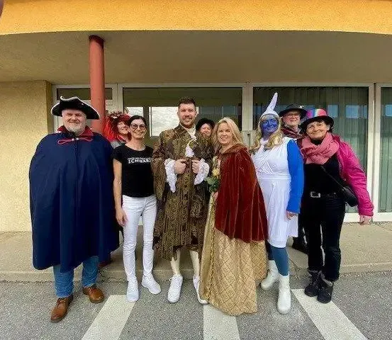 Eine Gruppe von Menschen in Kostümen steht vor einem Gebäude, einige tragen Hüte, Schuhe und Handschuhe. Eine Frau in einem roten Kleid hält einen Blumenstrauß.
