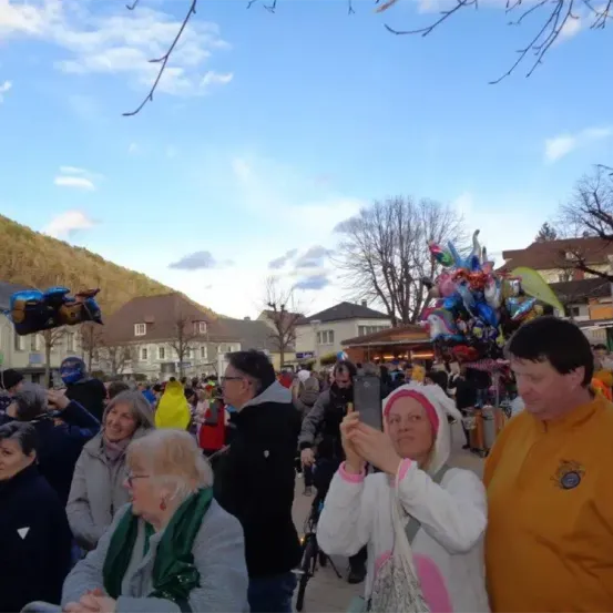 Eine Gruppe von Menschen versammelt sich im Freien, einige in Winterkleidung. Eine Frau in einer weißen Kapuzenjacke macht ein Foto mit ihrem Handy. In der Nähe beobachtet ein Mann in einem gelben Mantel. Im Hintergrund sind Gebäude, Bäume und ein Hügel zu sehen.