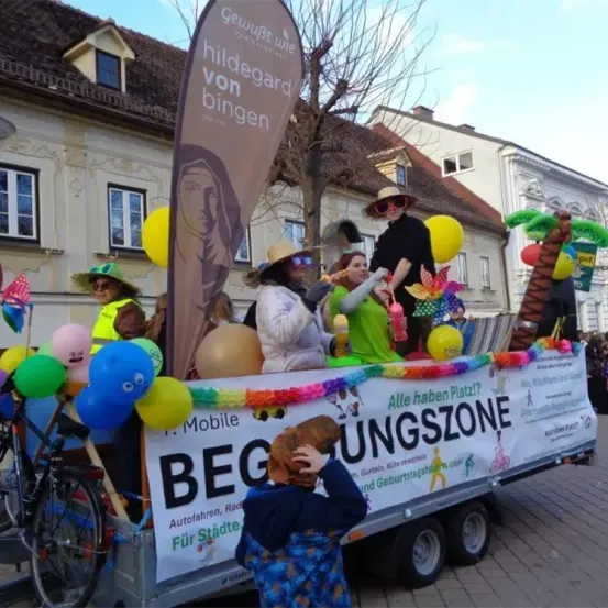 Ein Paradewagen mit einem Banner, das 'Alle haben Platz?' lautet, zeigt Menschen in Kostümen, Luftballons und einem Fahrrad. Ein Kind beobachtet von der Straße aus.