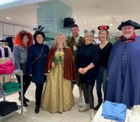 Eine Gruppe von Menschen in Kostümen posiert für ein Foto in einem Bekleidungsgeschäft, wobei einige mittelalterliche und Disney-inspirierte Outfits tragen.