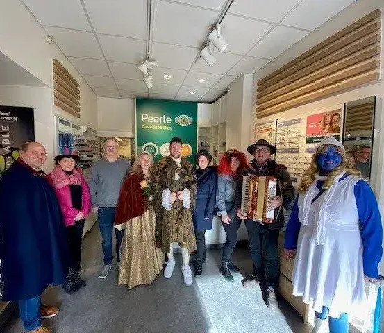 Eine Gruppe von Menschen in Kostümen posiert für ein Foto in einem Raum mit Regalen voller Brillen und einem grünen Banner mit dem Wort Pearle.