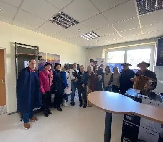 Eine Gruppe von Menschen in Winterkleidung steht in einem Raum mit einer Holztür und einem Fenster. Einige tragen Hüte, und eine Person spielt Akkordeon.