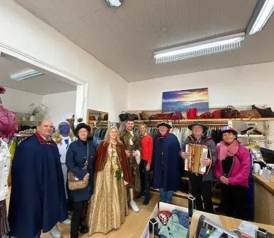 Eine Gruppe von Personen in historischen Gewändern steht in einem Bekleidungsgeschäft und lächelt für die Kamera. Ein Mannequin in einer weißen Jacke steht links, und Regale mit Handtaschen befinden sich dahinter.