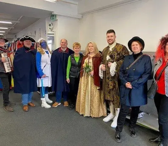 Eine Gruppe von Menschen in Kostümen steht in einem Raum, wobei eine Person ein Akkordeon spielt. Der Hintergrund scheint ein Geschäft zu sein, mit Kleiderständern im Hintergrund.