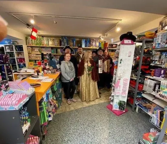 Eine Gruppe von Menschen, darunter Frauen in Kostümen, steht in einem Geschäft mit Regalen voller verschiedener Artikel und einer Theke im Hintergrund.