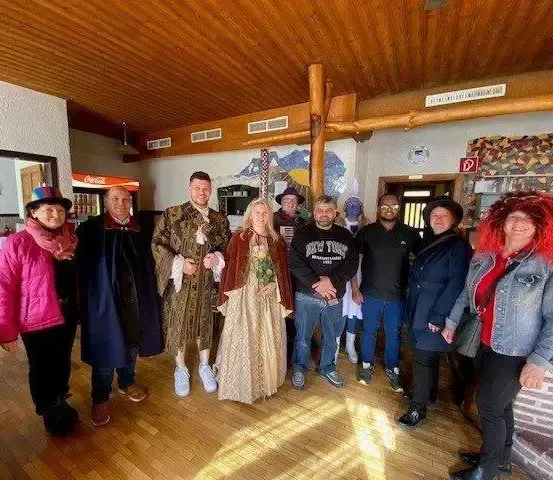 Eine Gruppe von Menschen in verschiedenen Kostümen, einschließlich einer Frau in einem historischen Kleid, steht in einem Raum mit Holzböden und einer Holzdecke. Hinter ihnen ist ein Tresen mit einer Ziegelwand und Regalen voller Gläser zu sehen.