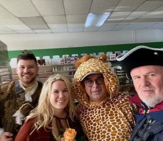 Eine Gruppe von Menschen posiert für ein Foto in einem Geschäft. Ein Mann in einem Giraffenkostüm, eine Frau in einer roten Jacke und ein Mann in einem Piratenhut. Hinter ihnen befinden sich Regale mit verschiedenen Artikeln.