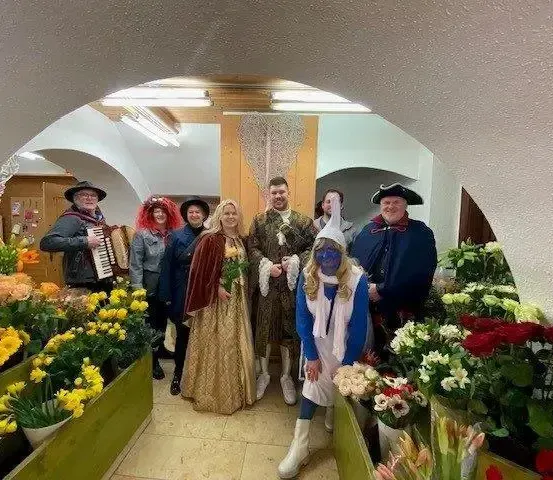 Eine Gruppe von Menschen in Kostümen posiert für ein Foto in einem Blumenladen, mit Blumen und einem Akkordeonspieler im Hintergrund.