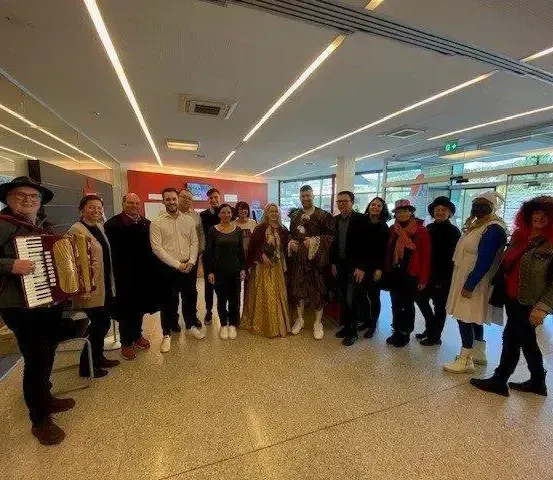 Eine Gruppe von Menschen in Kostümen posiert für ein Foto in einem Saal mit roter Wand und weißen Deckenleuchten. Ein Mann spielt Akkordeon.