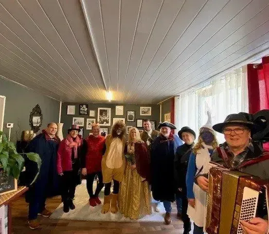 Eine Gruppe von Menschen steht in einem Raum, einige in Kostümen gekleidet. Ein Mann spielt Akkordeon. Der Raum hat eine weiße Decke und grüne Wände. Es gibt Bilderrahmen und einen Topf mit Pflanzen.