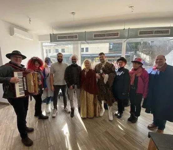 Eine Gruppe von Menschen in Kostümen und Hüten steht zusammen in einem Raum mit Holzboden, ein Mann spielt Akkordeon, und ein Fenster zeigt die Außenseite.