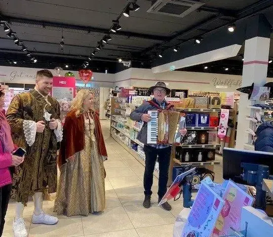 Eine Gruppe von Menschen ist in einem Geschäft. Ein Mann spielt Akkordeon, während andere in Kostümen gekleidet sind. Eine Frau hält ein Telefon.