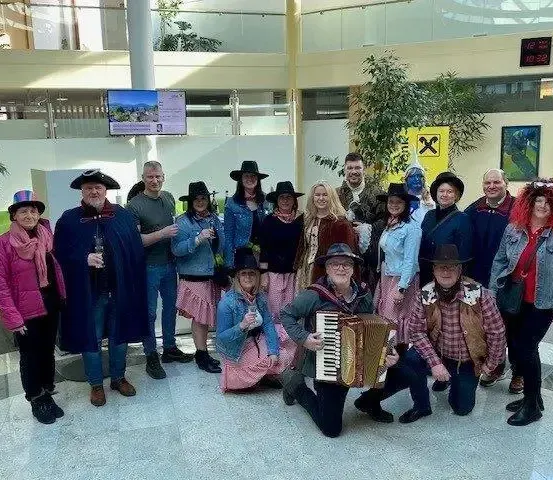 Eine Gruppe von Menschen in verschiedenen Kostümen posiert für ein Foto in einem Gebäude. Einige tragen Hüte, während andere Instrumente spielen. Ein gelbes Schild und ein Monitor sind im Hintergrund zu sehen.