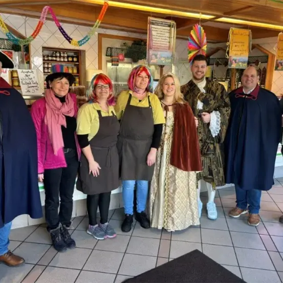 Eine Gruppe von Menschen in Kostümen posiert für ein Foto in einem Restaurant. Einige tragen Schürzen und andere Umhänge. Dahinter befinden sich Regale mit Flaschen und Gläsern, und ein Schild ist an der Wand angebracht.