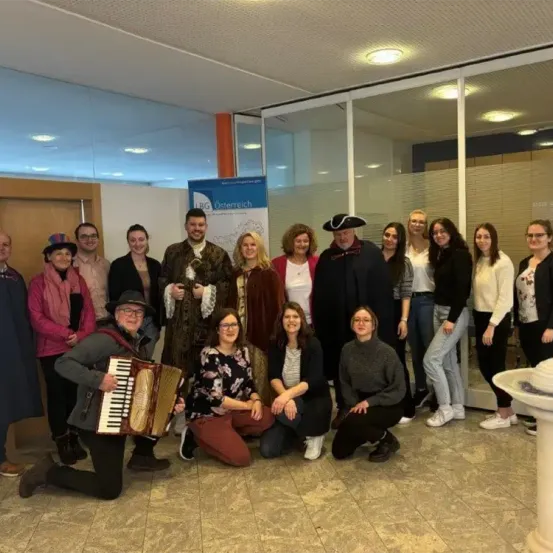 Eine Gruppe von Menschen in historischen Kostümen posiert für ein Foto in einem Raum mit Glaswänden. Ein Banner im Hintergrund trägt die Aufschrift 'LBG Osterreich'.