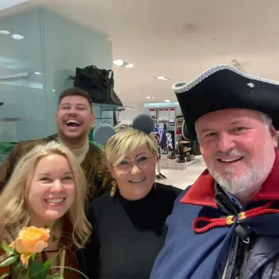 Vier Personen lächeln für ein Foto. Ein Mann in einem Piratenhut steht neben einer Frau mit einer Rose. Dahinter lächeln ein Mann in einem Kostüm und eine weitere Frau. Ein Handtasche hängt an der Wand im Hintergrund.