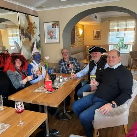 Fünf Personen sitzen um einen Tisch in einem Restaurant, alle in Kostümen. Sie halten Getränke in der Hand und wirken fröhlich. Ein Mann trägt einen Piratenhut.