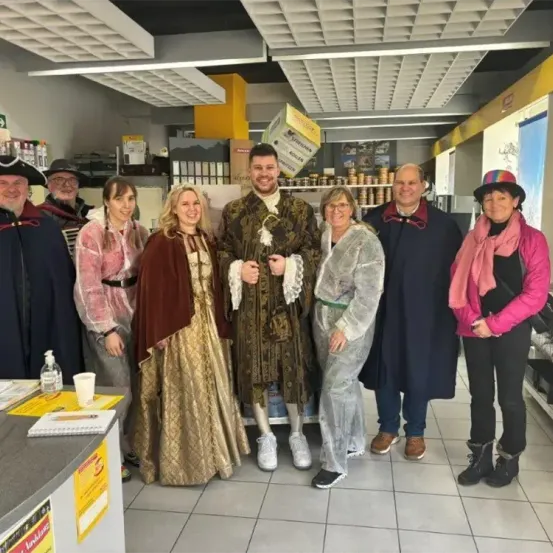 Eine Gruppe von Menschen in historischen Kostümen steht in einem Geschäft. Ein Mann in der Mitte trägt ein goldfarbenes Outfit. Die Menschen lächeln für ein Foto.