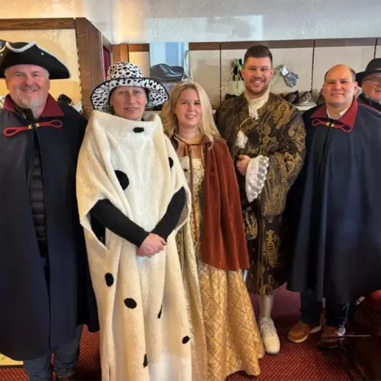 Fünf Menschen posieren für ein Foto in einem Raum. Sie tragen alle Kostüme. Der Mann auf der linken Seite trägt einen Piratenhut und eine rote Schleife um den Hals. Die Frau neben ihm trägt einen weißen Umhang mit schwarzen Punkten. Die Frau in der Mitte trägt ein goldenes Kleid. Der Mann auf der rechten Seite trägt einen braunen Umhang.