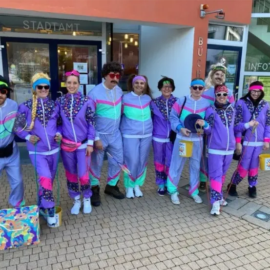 Eine Gruppe Erwachsener, gekleidet in leuchtenden Trainingsanzüge im Stil der 1980er Jahre, posiert für ein Foto vor einem Gebäude. Jeder hält einen Eimer.