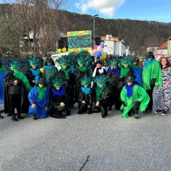Eine Gruppe von Menschen in bunten Kostümen posiert für ein Foto auf der Straße. Hinter ihnen wird ein Banner mit den Buchstaben 'NFG' gezeigt.