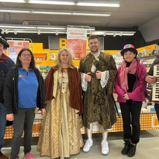 Eine Gruppe von Erwachsenen in Kostümen posiert für ein Foto in einer Buchhandlung. Ein Mann in der Mitte trägt ein historisches Kostüm. Hinter ihnen steht ein Plakat mit der Aufschrift 'DAS BESTE VON #BOOKTOK!'.