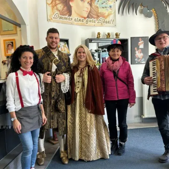 Fünf Menschen in Kostümen, zwei Männer und drei Frauen, posieren für ein Foto. Ein Mann spielt Akkordeon, während andere lächeln. Ein Banner mit einem Bild einer Frau ist über ihnen.