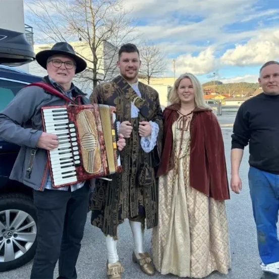 Vier Personen stehen auf einem Parkplatz, zwei Männer und zwei Frauen. Ein Mann spielt Akkordeon, der andere trägt ein historisches Kostüm. Die Frauen tragen traditionelle Kleider. Ein Auto ist hinter ihnen geparkt.