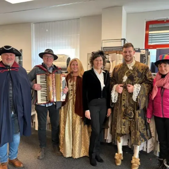 Eine Gruppe von Menschen in Kostümen, darunter ein Mann, der ein Akkordeon spielt, steht in einem Raum mit einer weißen Wand.