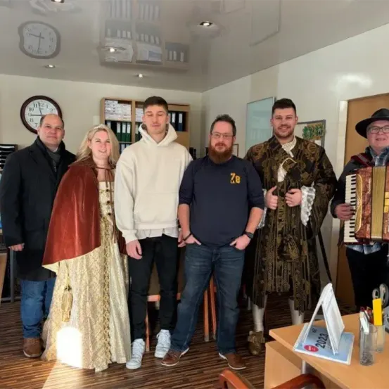 Eine Gruppe von Menschen steht in einem Raum, gekleidet in verschiedene Kostüme. Ein Mann in einer weißen Kapuzenjacke und Jeans steht neben einem Mann in einem blauen T-Shirt. Daneben posiert eine Frau in einem mittelalterlichen Kleid. Ein weiterer Mann in einem Kostüm spielt Akkordeon. Hinter ihnen sind eine Uhr und ein Regal mit Büchern zu sehen.