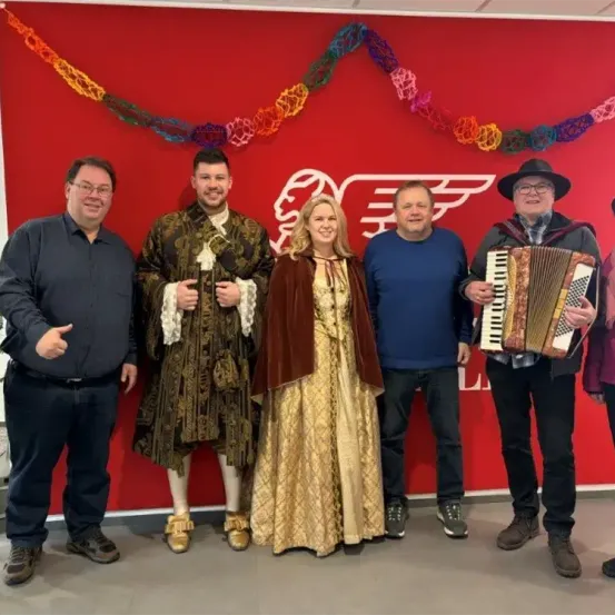Eine Gruppe von Menschen in Kostümen steht vor einer roten Wand. Eine Frau in einem goldenen Kleid, ein Mann in einem schwarzen Kostüm und drei Männer in Freizeitkleidung posieren für ein Foto.