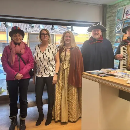 Fünf Personen stehen im Innenraum. Drei Frauen und zwei Männer. Der Mann auf der rechten Seite spielt ein Akkordeon. Hinter ihnen befindet sich eine Glaswand mit Büchern auf der rechten Seite.