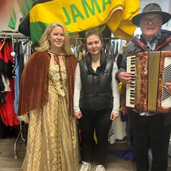 Drei Personen stehen in einem Bekleidungsgeschäft. Die Frau in der Mitte trägt eine schwarze Weste. Die Frau auf der linken Seite trägt ein goldenes Kleid und einen Umhang. Der Mann auf der rechten Seite hält ein Akkordeon. Hinter ihnen hängen Kleidungsstücke an einem Ständer.