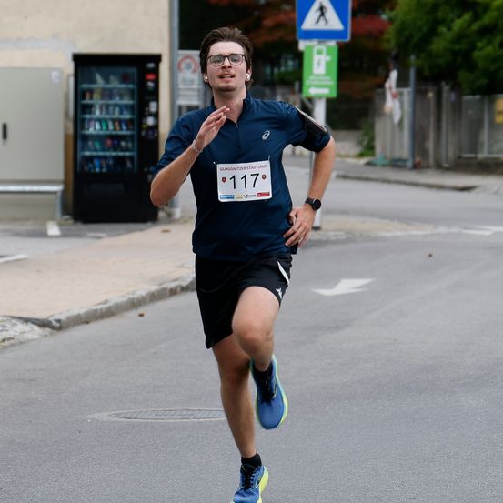 Ein Mann mit der Nummer 117 auf dem Shirt läuft einen Marathon auf der Straße. Hinter ihm steht ein Verkaufsautomat und ein Verkehrsschild.