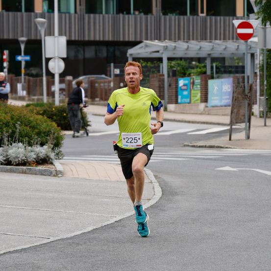Ein Läufer mit der Nummer 225 auf seinem Shirt läuft auf einer Straße. Hinter ihm geht eine Person mit einem Fahrrad. Im Hintergrund befinden sich Gebäude, Pflanzen und Schilder.