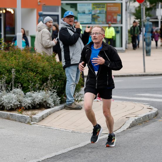 Ein älterer Mann läuft an der Straßenecke, trägt ein blaues Shirt und schwarze Shorts. Hinter ihm beobachtet ein Mann in einer grauen Jacke und Jeans. Im Hintergrund ist eine Frau in einem hellblauen Shirt zu sehen.
