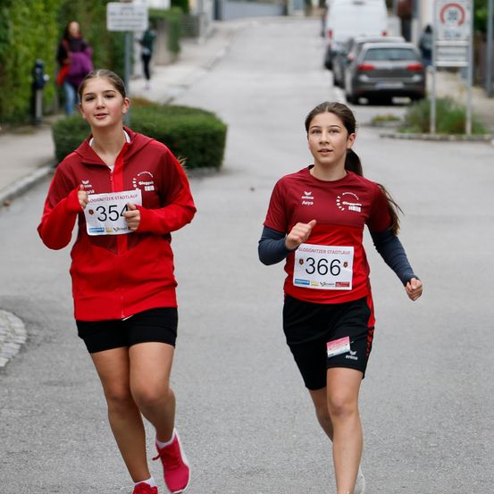 Zwei junge Frauen in roter und schwarzer Sportkleidung mit Nummernschildern laufen auf einer Straße mit geparkten Autos und Schildern am Straßenrand einen Marathon.