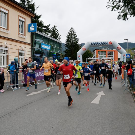 Eine Gruppe von Läufern nimmt an einem Marathon auf einer Straße teil, mit Zuschauern. Der Eingang ist durch einen 'Sparkasse'-Bogen gekennzeichnet. Der Himmel ist bedeckt.