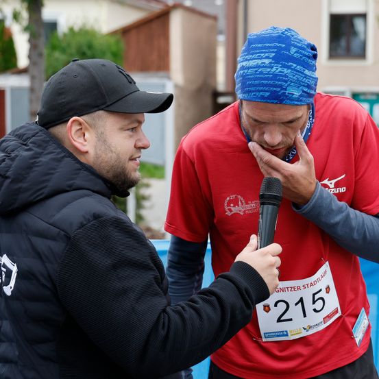 Ein Marathonläufer wird von einem Reporter interviewt und trägt ein rotes Shirt mit der Nummer 215. Er hat eine blaue Kopfbedeckung und hält ein Mikrofon.