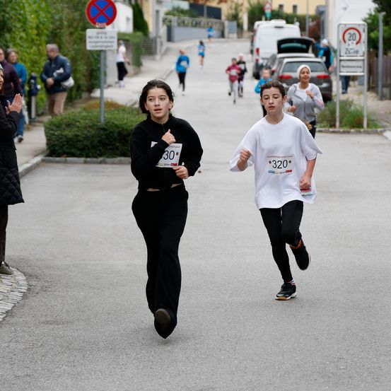 Zwei junge Läufer nehmen an einem Rennen auf einer asphaltierten Straße teil. Das Mädchen trägt ein schwarzes Shirt und Hose, der Junge ein weißes Shirt. Zuschauer stehen am Straßenrand, und der Verkehr wird durch Straßenschilder geregelt.