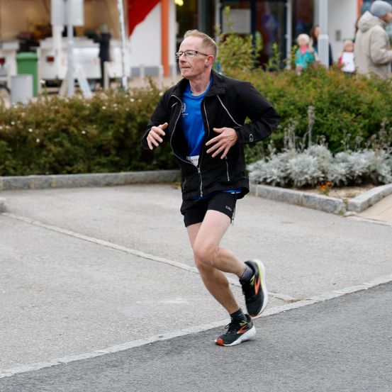 Ein älterer Mann läuft auf einer Straße, trägt eine Brille, eine schwarze Jacke, ein blaues T-Shirt, schwarze Shorts und Turnschuhe. Hinter ihm stehen Büsche und Menschen in der Nähe eines Gebäudes.