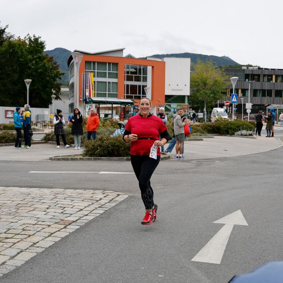 Eine Frau läuft in einem Marathon, trägt ein rotes Shirt und schwarze Hose. Zuschauer beobachten sie vom Straßenrand, mit Gebäuden und Bäumen im Hintergrund.