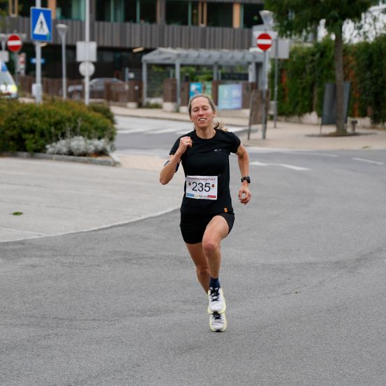 Eine Frau in einem schwarzen Shirt mit der Nummer 235 läuft auf einer Straße. Im Hintergrund befinden sich Büsche und Gebäude.